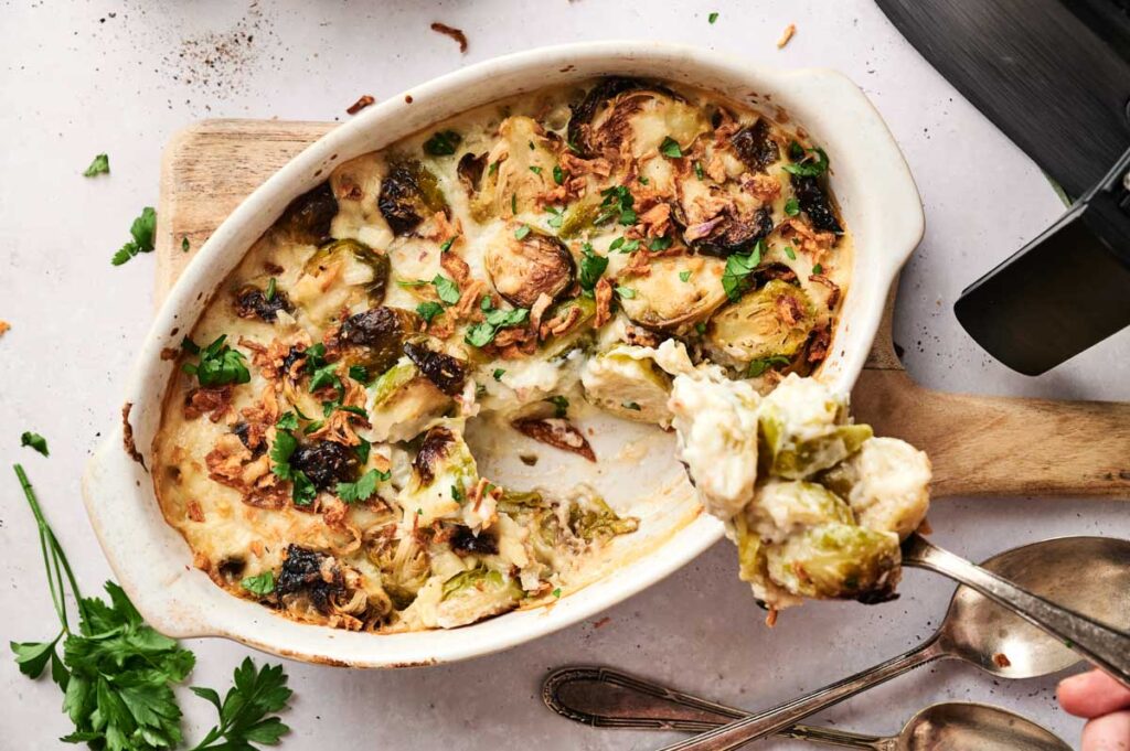 A baked creamy Air Fryer Brussels Sprouts Casserole sits in a white dish, with a serving spoon lifting a portion. Fresh parsley and two spoons are on the side.