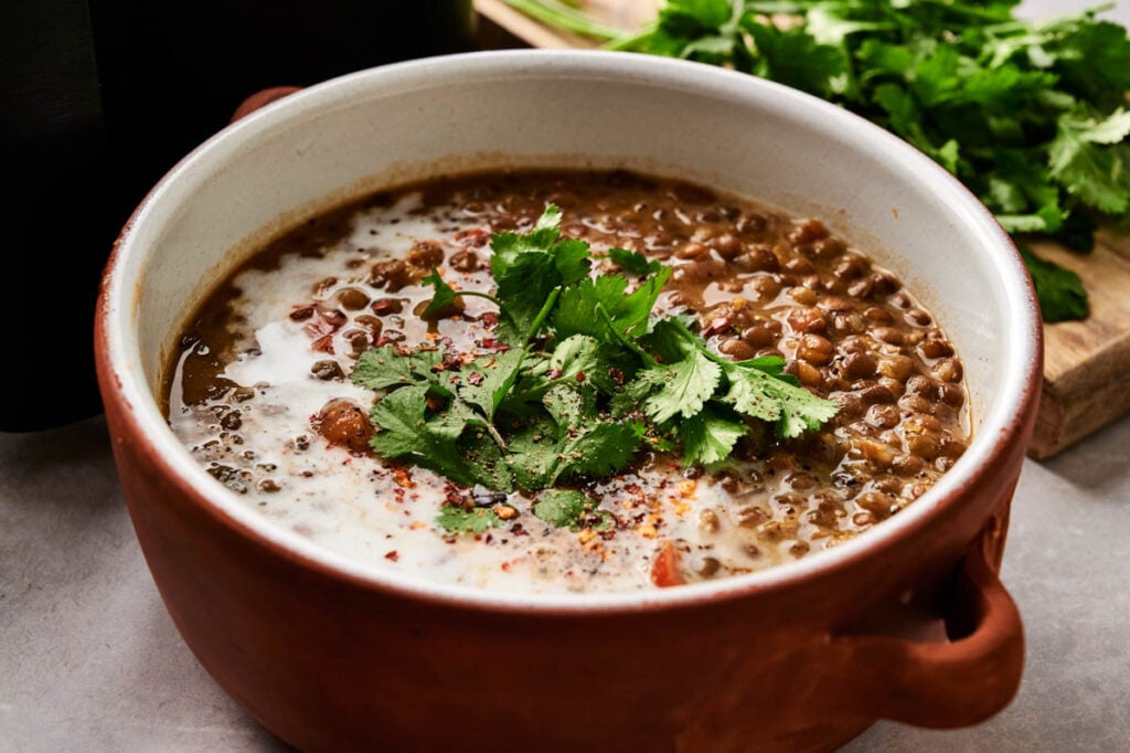 A bowl of Air Fryer Lentil Soup topped with a swirl of cream and fresh cilantro, with extra cilantro on a wooden board in the background.