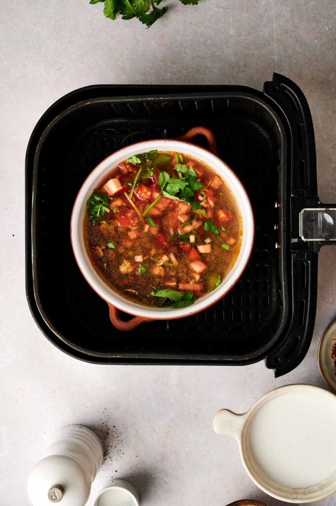 A bowl of Air Fryer Lentil Soup with chopped vegetables and herbs sits inside an air fryer basket on a light-colored countertop.