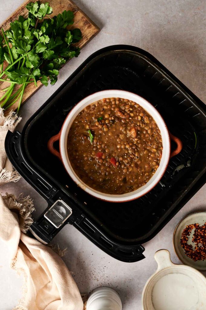 A bowl of Air Fryer Lentil Soup sits inside an air fryer basket, surrounded by fresh cilantro on a cutting board and small bowls of spices on a light countertop.