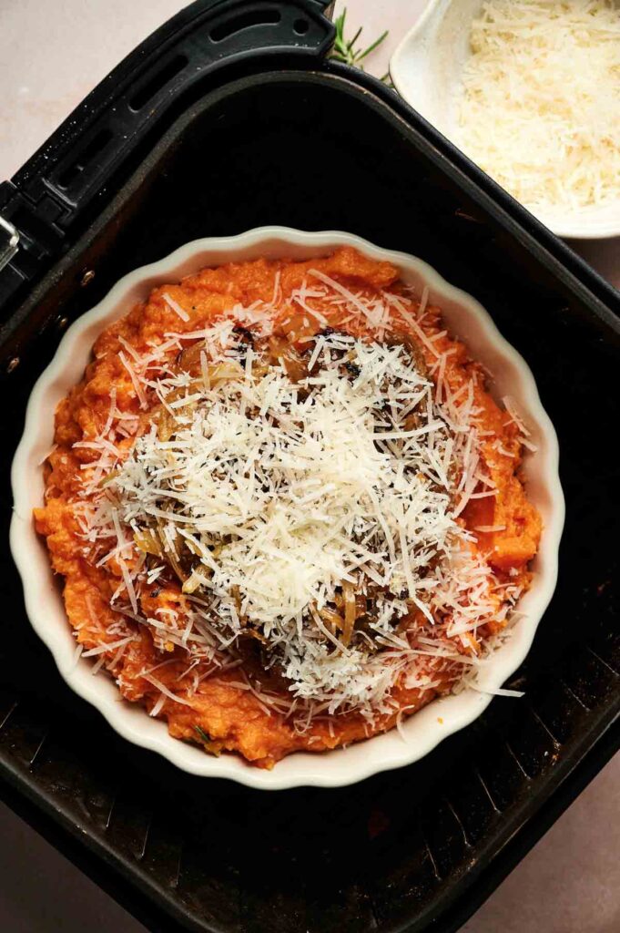 A round baking dish with a tomato-based casserole topped with shredded cheese and herbs sits in an air fryer basket. Nearby, a bowl of grated cheese and a side of Air Fryer Mashed Sweet Potatoes complete the scene.
