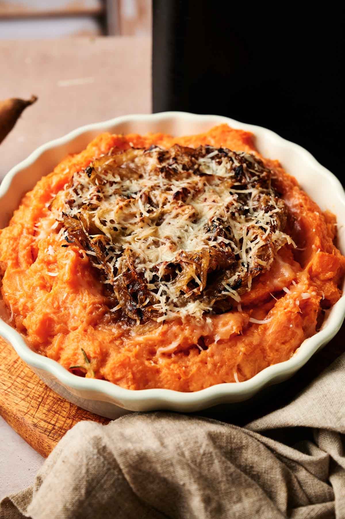 A round dish filled with Air Fryer Mashed Sweet Potatoes, topped with crispy caramelized onions and grated cheese, placed on a wooden board.