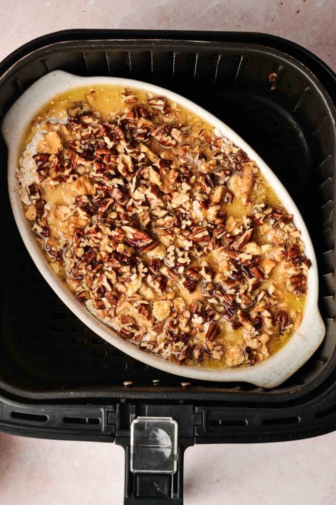 Oval dish with chopped pecans and a baked Air Fryer Pumpkin Dump Cake inside, placed in an air fryer basket on a light surface.