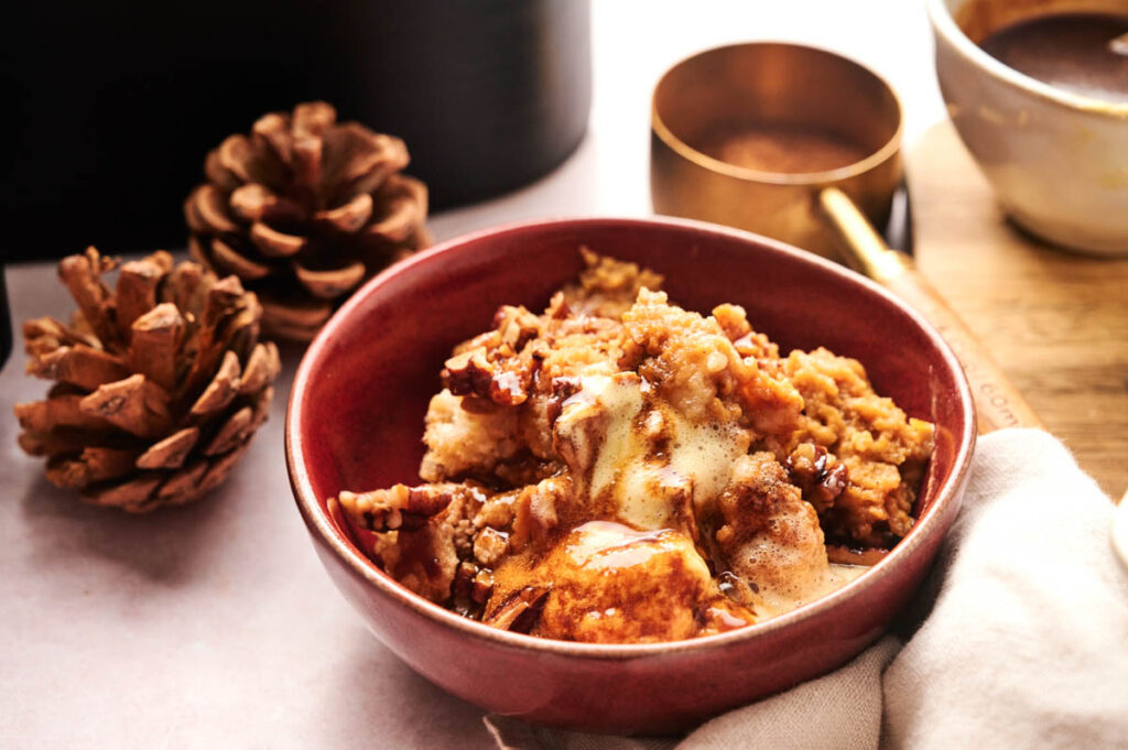 A red bowl filled with Air Fryer Pumpkin Dump Cake, topped with ice cream, sauce, and pecans, is placed next to pine cones, a brass cup, and a mug.