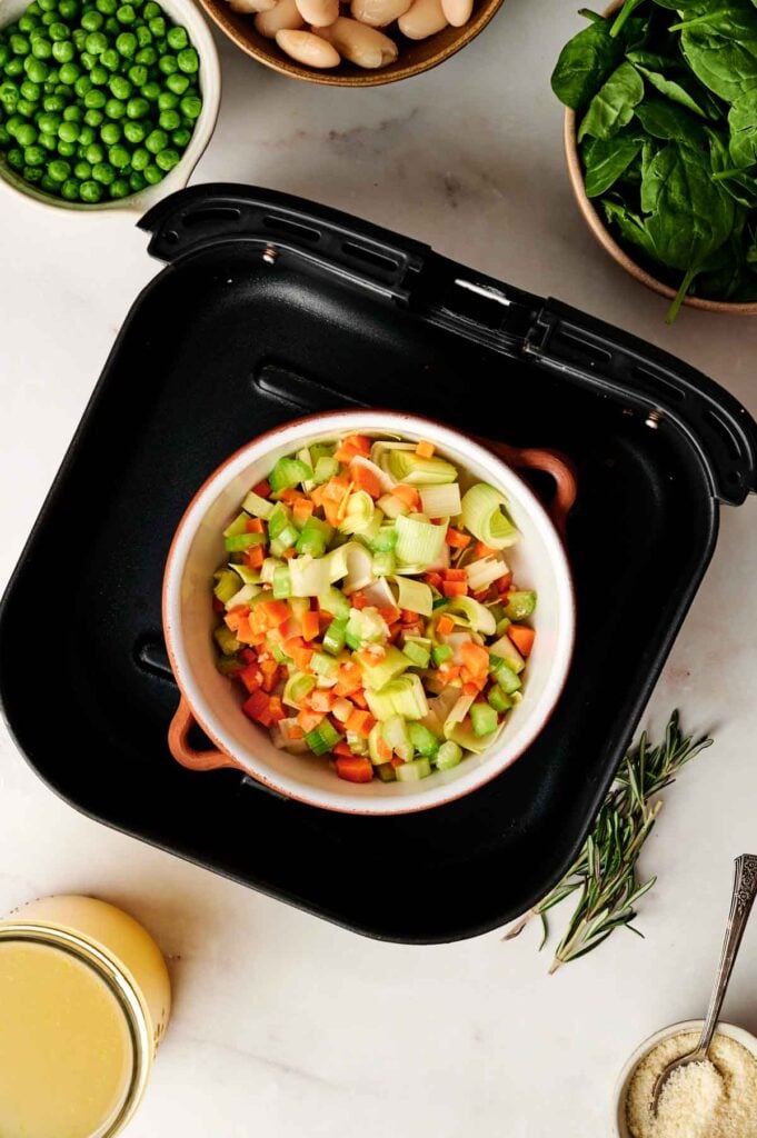 Chopped carrots, celery, and leeks in a dish inside an air fryer, surrounded by bowls of peas, spinach, white beans, broth, and seasonings on a white countertop—everything you need to prepare a hearty Air Fryer Vegetable Soup.