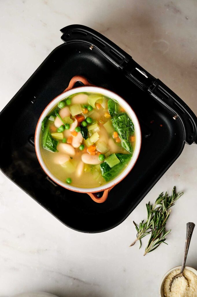 A pot of Air Fryer Vegetable Soup with beans, peas, carrots, and leafy greens sits inside an air fryer basket on a white surface, with rosemary and grated cheese nearby.