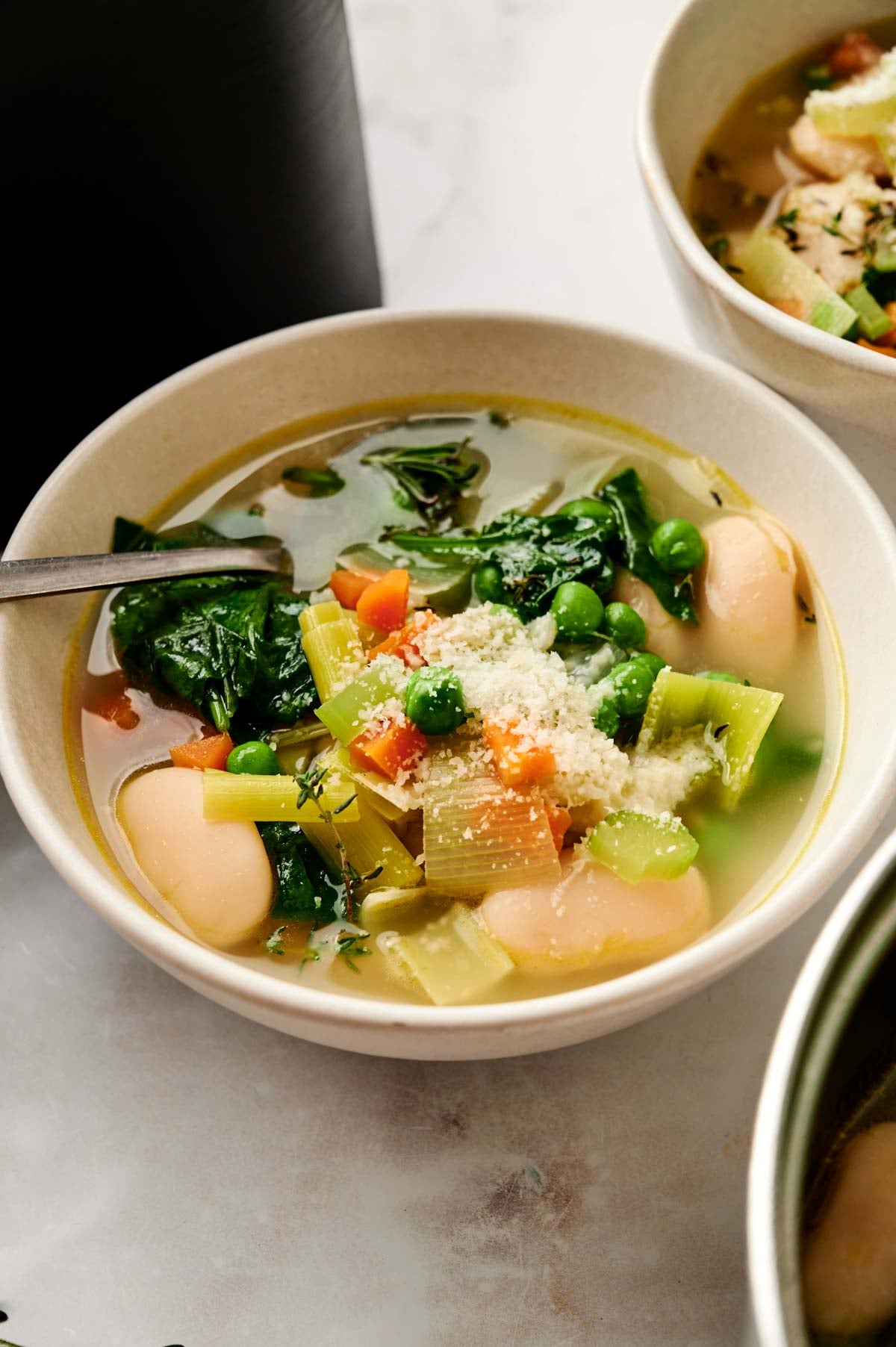 A bowl of Air Fryer Vegetable Soup with spinach, celery, peas, carrots, beans, and grated cheese on top, with a spoon resting inside the bowl.