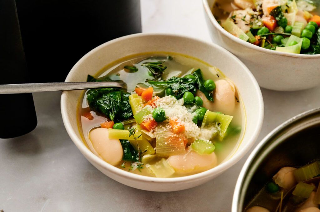 A bowl of Air Fryer Vegetable Soup with spinach, carrots, peas, celery, and white beans, topped with grated cheese and served with a spoon.