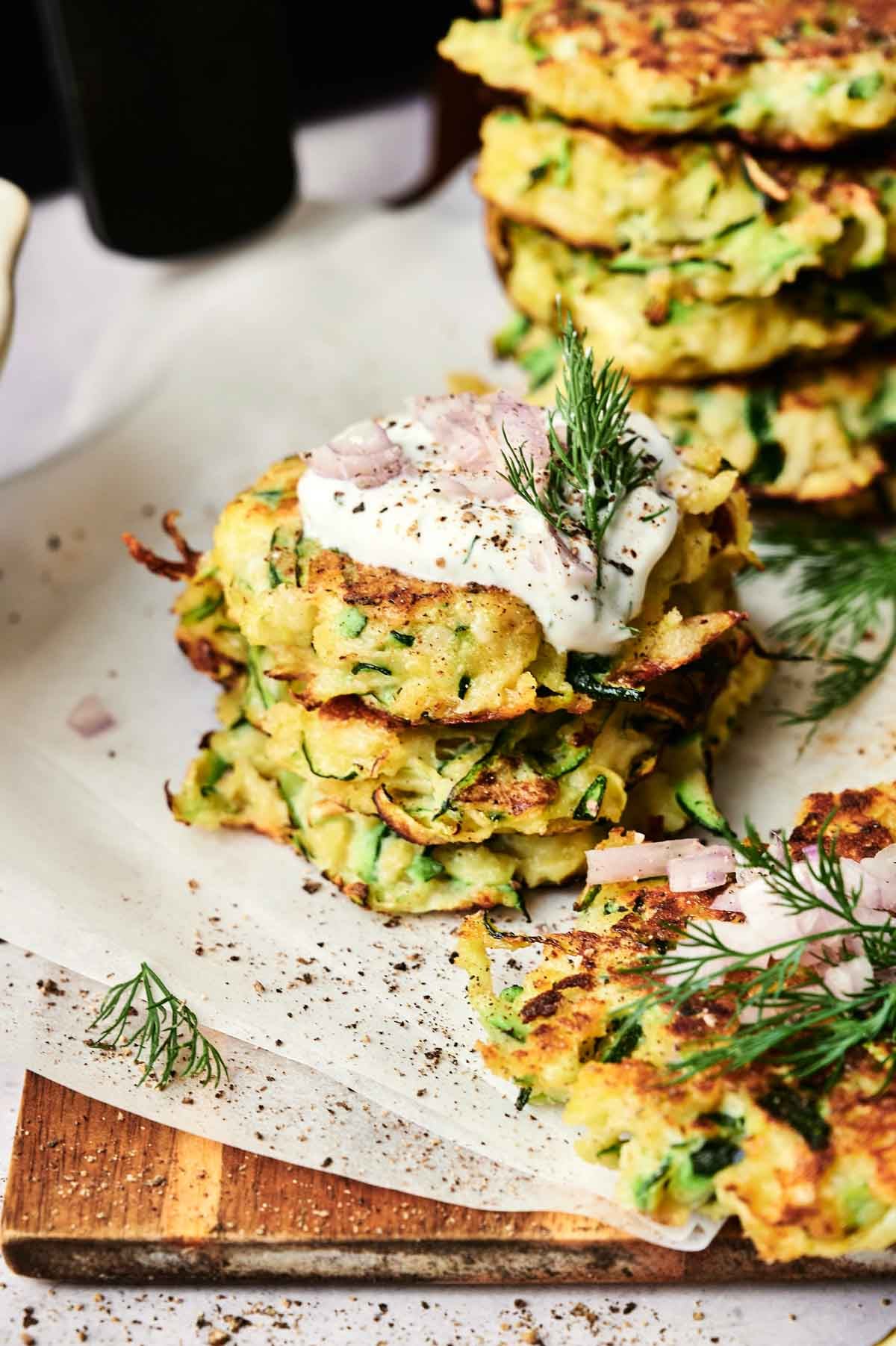 A stack of Air Fryer Zucchini Fritters topped with sour cream, chopped onions, fresh dill, and black pepper, with more fritters in the background.