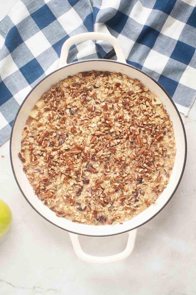 A white baking dish filled with an unbaked Baked Oatmeal and nut mixture sits on a white surface next to a blue and white checkered cloth.