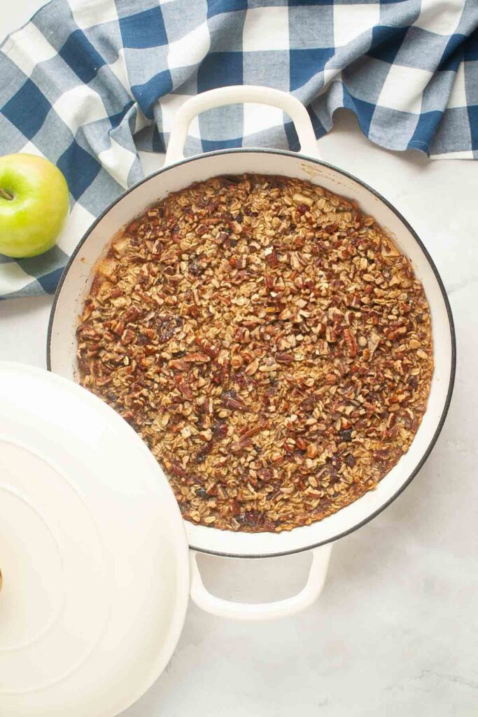 A round white baking dish filled with a delicious baked oatmeal and nut mixture sits on a marble surface next to a green apple and a blue and white checkered cloth.