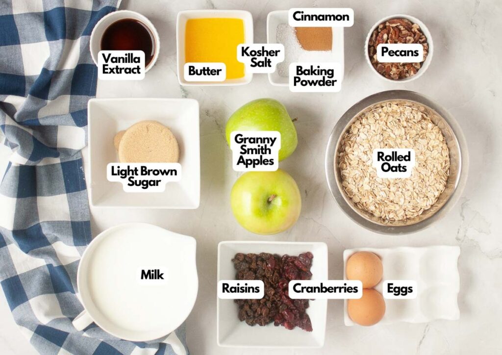 Top-down view of labeled Baked Oatmeal ingredients: vanilla extract, butter, kosher salt, cinnamon, baking powder, pecans, oats, eggs, cranberries, raisins, milk, brown sugar, and apples.