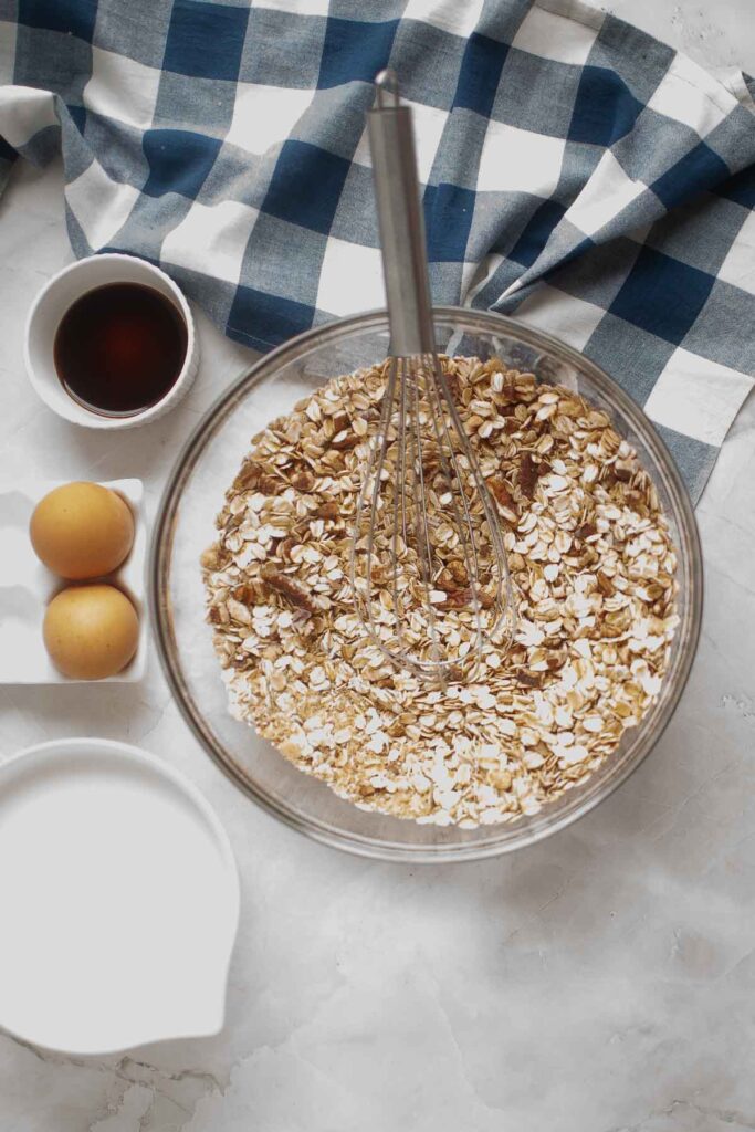 A glass bowl with oats and a whisk, beside two eggs, a cup of vanilla extract, a cup of milk, and a blue-and-white checkered cloth on a marble surface—everything you need to prepare delicious baked oatmeal.