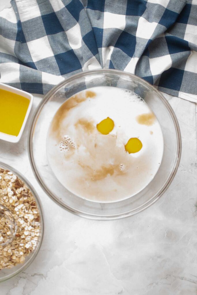 A glass bowl with eggs, milk, and vanilla—perfect for preparing baked oatmeal—sits next to a dish of melted butter, a bowl of oats, and a blue-and-white checkered cloth on a marble surface.
