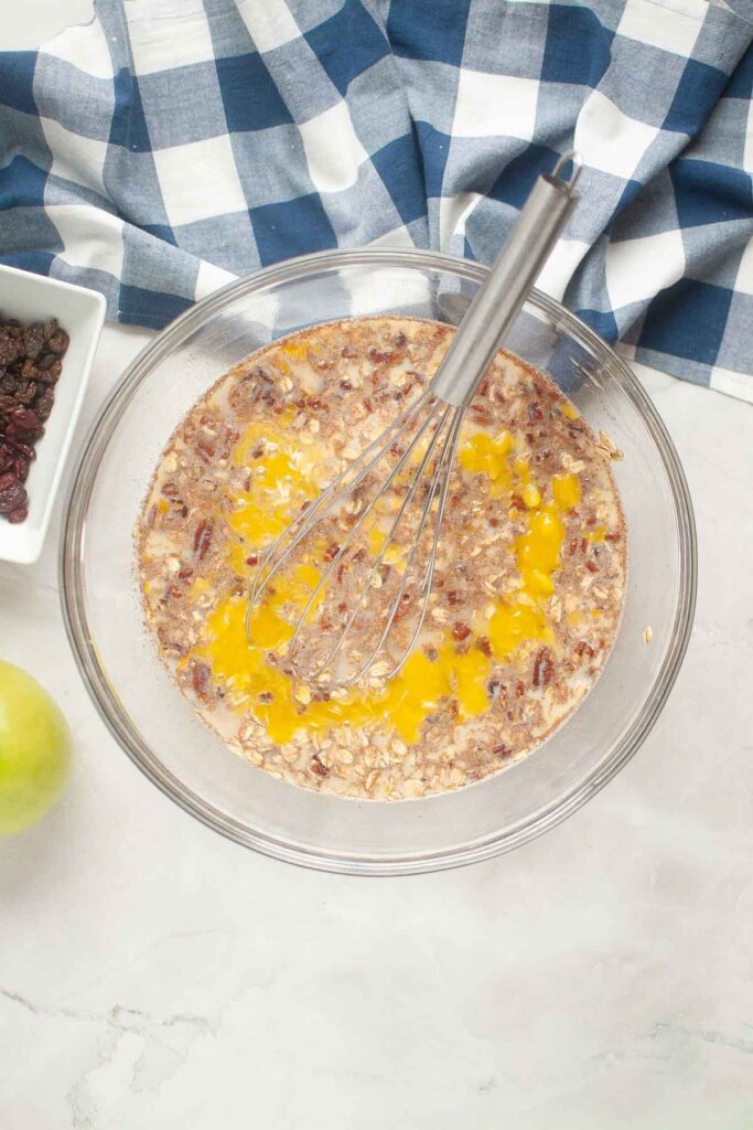 A glass bowl with ingredients for baked oatmeal—oatmeal, raisins, milk, and eggs—being mixed with a whisk, next to a green apple and a blue checkered cloth.