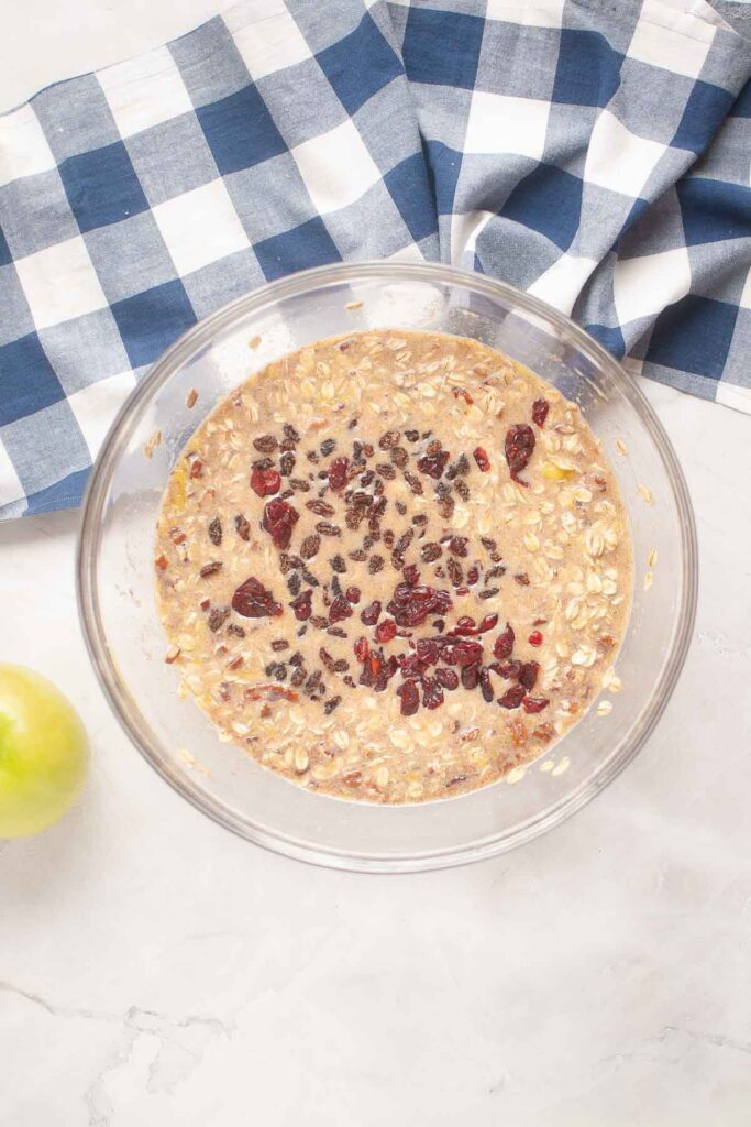 A glass bowl filled with Baked Oatmeal batter, topped with raisins and dried cranberries, sits on a white surface next to a blue plaid cloth and a green apple.