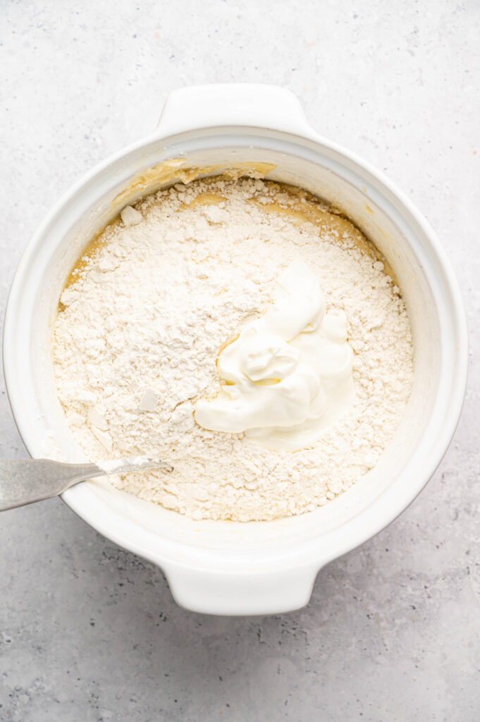 A white mixing bowl containing flour and a dollop of sour cream on top of a yellow batter for Boston Cream Pie Cupcakes, with a metal spoon partially visible on the left side.
