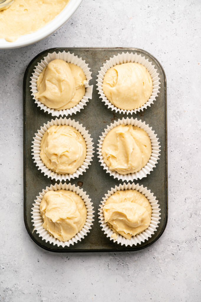A muffin tin with six paper liners, each filled with unbaked Boston Cream Pie Cupcake batter, sits on a light-colored countertop.