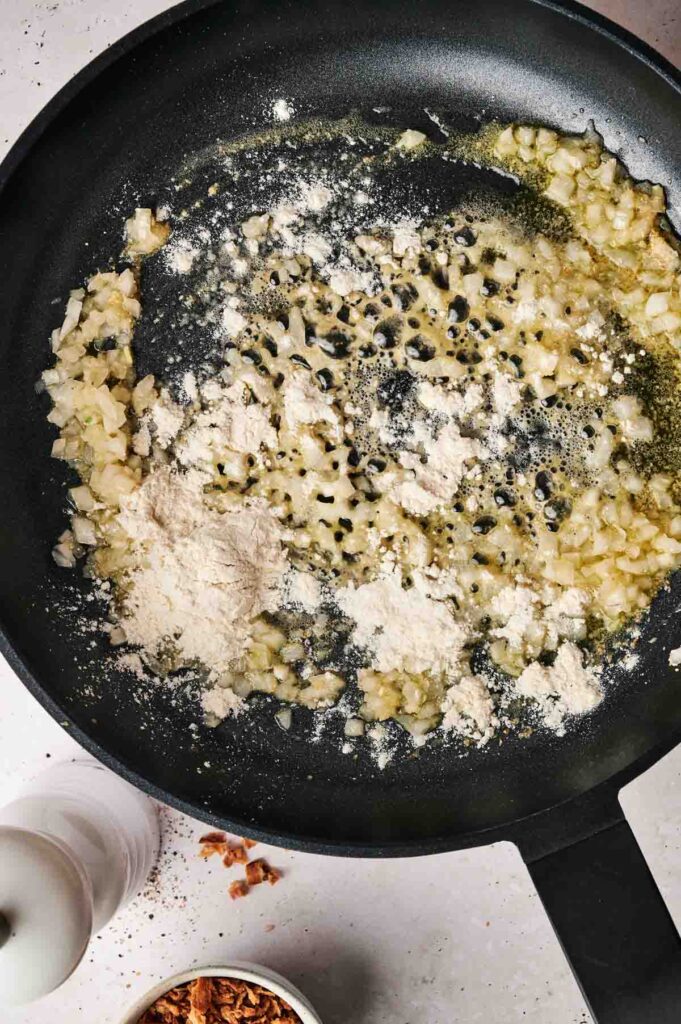 Chopped onions sautéing in butter in a skillet, with flour sprinkled on top, next to a pepper grinder and a small bowl of fried onions—perfect ingredients for pairing with an Air Fryer Brussels Sprouts Casserole.