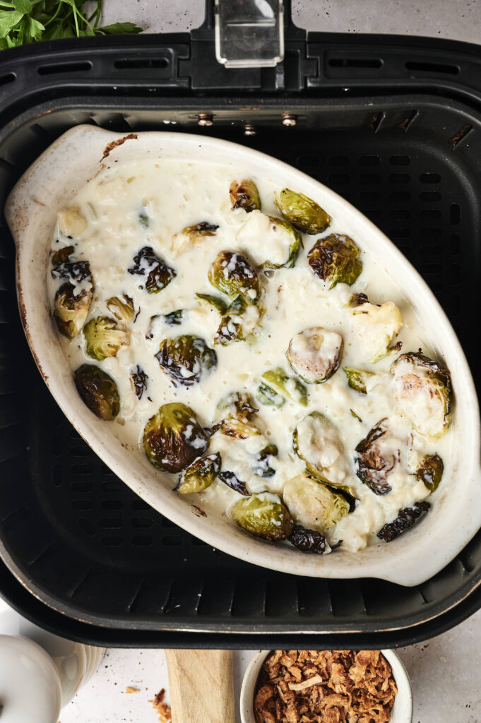 Oval dish with Air Fryer Brussels Sprouts Casserole in creamy white sauce, placed inside an air fryer basket. Crispy fried onions and fresh herbs are visible nearby on the countertop.
