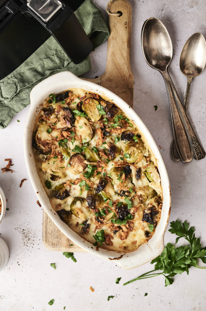 A baked casserole featuring roasted Brussels sprouts, inspired by an Air Fryer Brussels Sprouts Casserole, topped with chopped herbs in a white oval dish on a wooden board, next to two spoons and fresh parsley on a light surface.