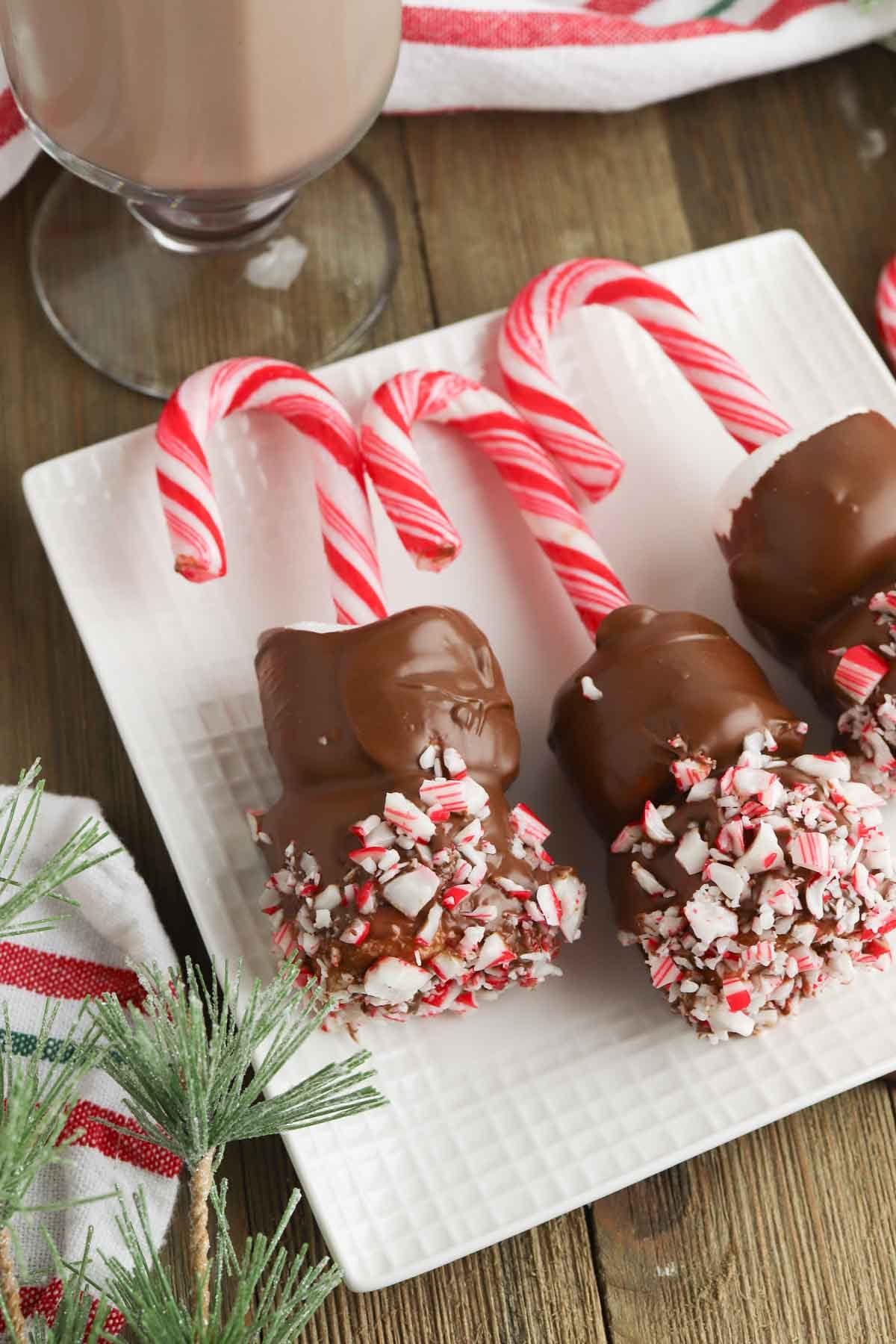 Three Candy Cane Hot Chocolate Stirrers&mdash;marshmallows dipped in chocolate and crushed peppermint on candy cane sticks&mdash;are arranged on a white square plate with a glass of chocolate milk nearby.