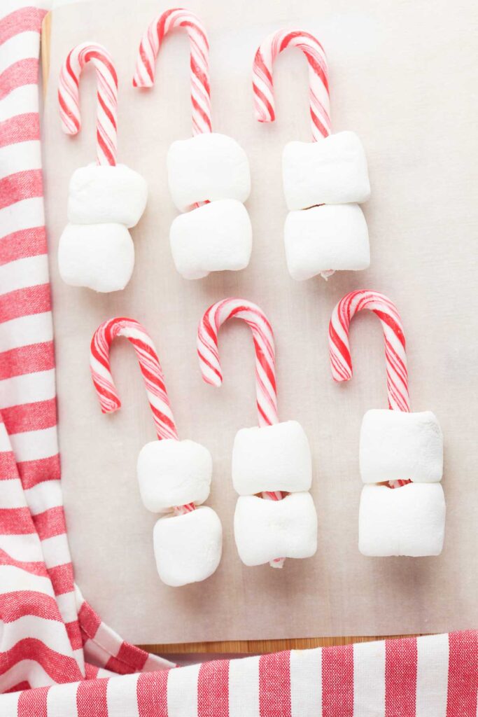 Peppermint candy canes threaded with fluffy marshmallows sit in two rows, ready to be used as festive stirrers for hot chocolate.