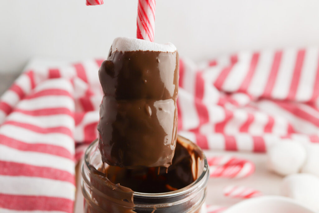 A marshmallow on a candy cane stick being dipped into melted chocolate, with a red and white striped cloth in the background, creating festive Candy Cane Hot Chocolate Stirrers.
