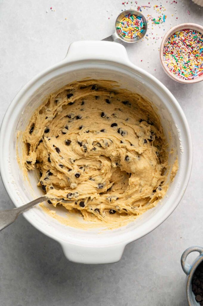 A white mixing bowl filled with chocolate chip cookie dough for a delicious Cookie Cake, a metal spoon, and bowls of colorful sprinkles on a light countertop.