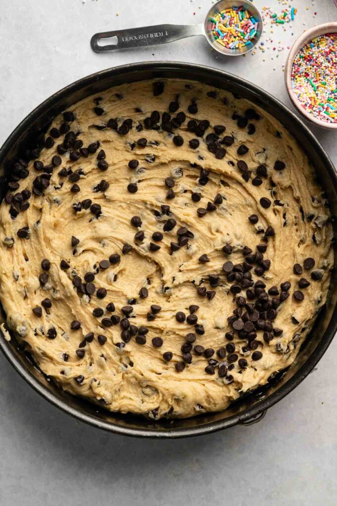 A round pan filled with raw chocolate chip cookie dough, perfect for baking a gooey cookie cake. Extra chocolate chips top the dough, while a measuring spoon and bowls of colorful sprinkles sit nearby on the counter.