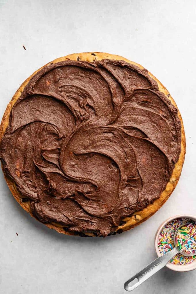 A large round Cookie Cake with chocolate frosting on top sits next to a measuring cup filled with colorful sprinkles on a light surface.