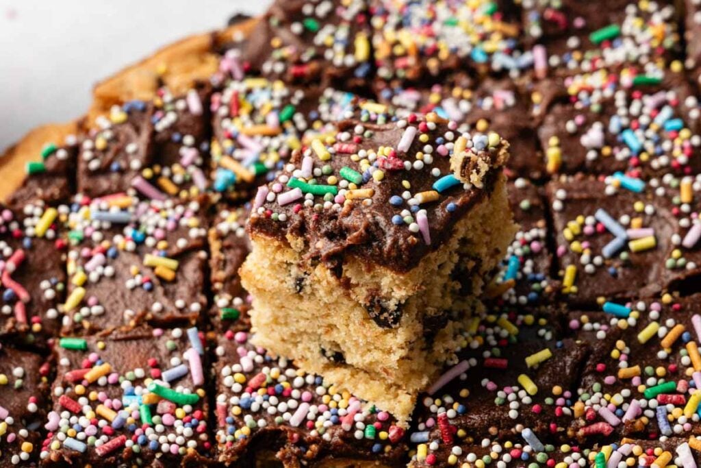 A tray of chocolate-frosted cookie cake squares with colorful sprinkles, with one piece lifted to show the inside of the cake.