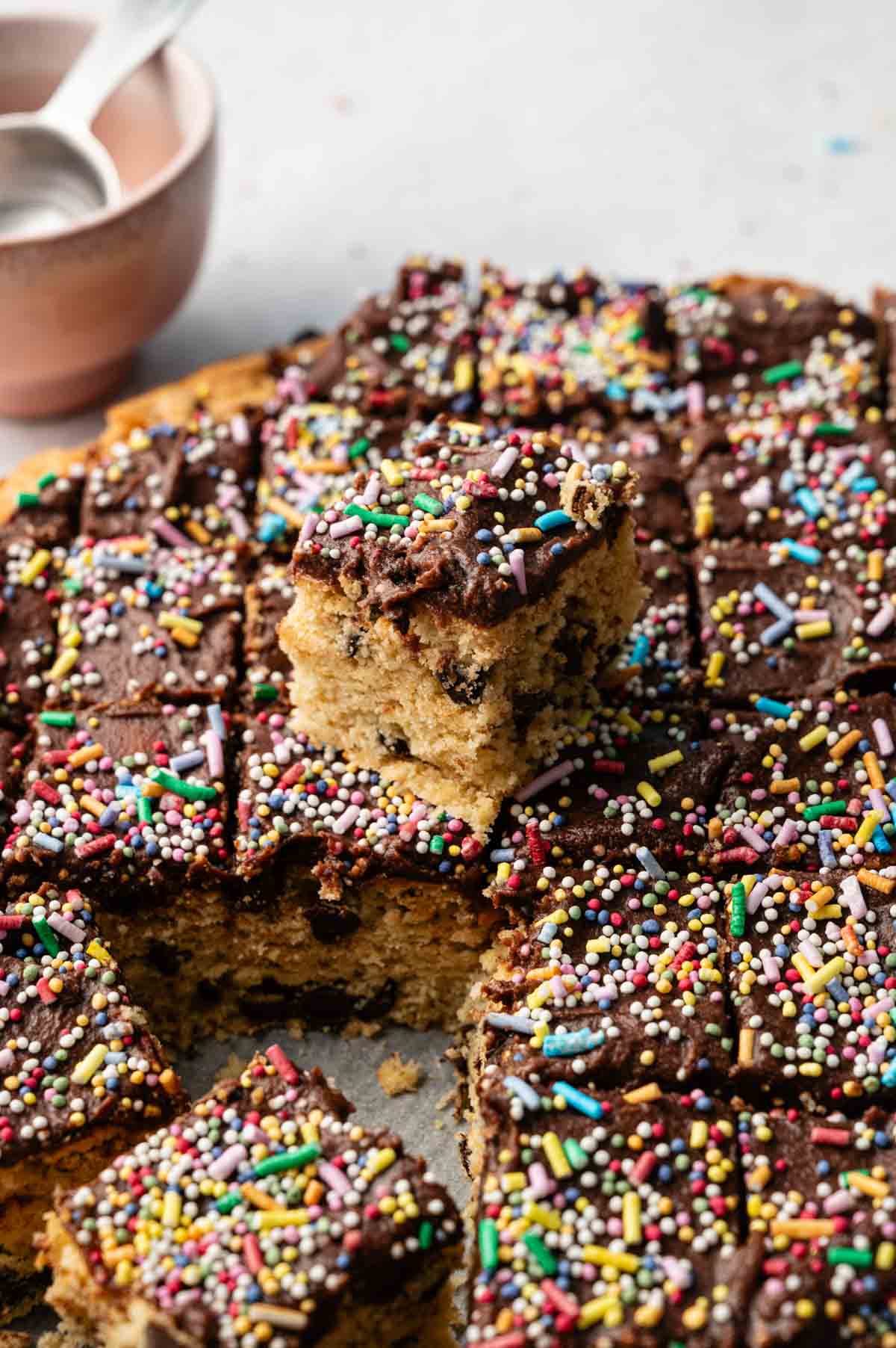 A chocolate-frosted, sprinkle-topped Cookie Cake sheet cut into squares with one piece placed on top, and a bowl with spoons in the background.