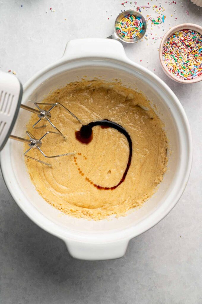 A mixing bowl with creamed batter, electric mixer beaters, and a swirl of vanilla extract on top—perfect for whipping up a delicious Cookie Cake; bowls of colorful sprinkles nearby.