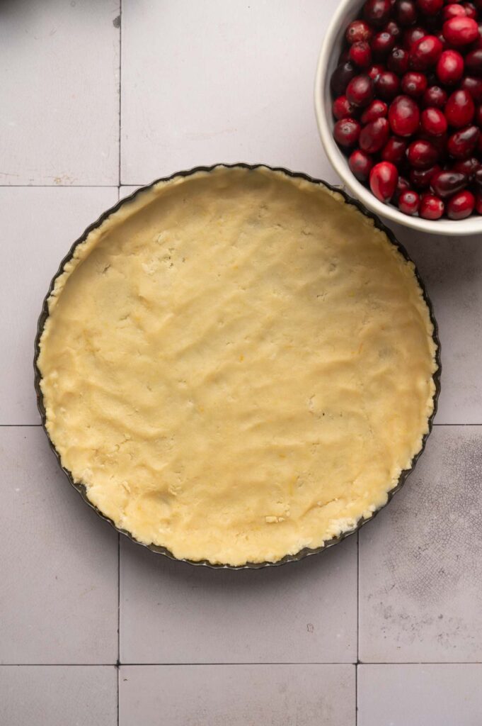 Unbaked pie crust in a round pan sits next to a white bowl filled with fresh cranberries, ready to be transformed into a delicious Cranberry Curd Tart on a tiled surface.