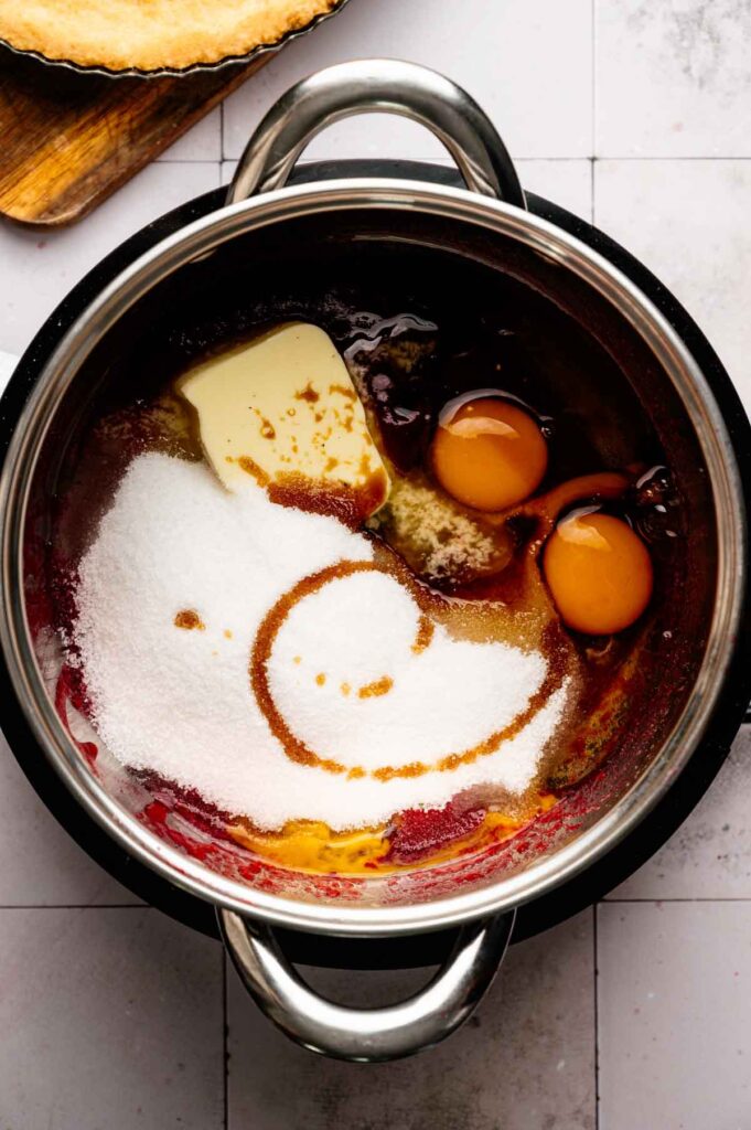 A saucepan containing butter, sugar, eggs, vanilla, and syrup—just the beginning for a luscious Cranberry Curd Tart—with a pie crust on a wooden board nearby.