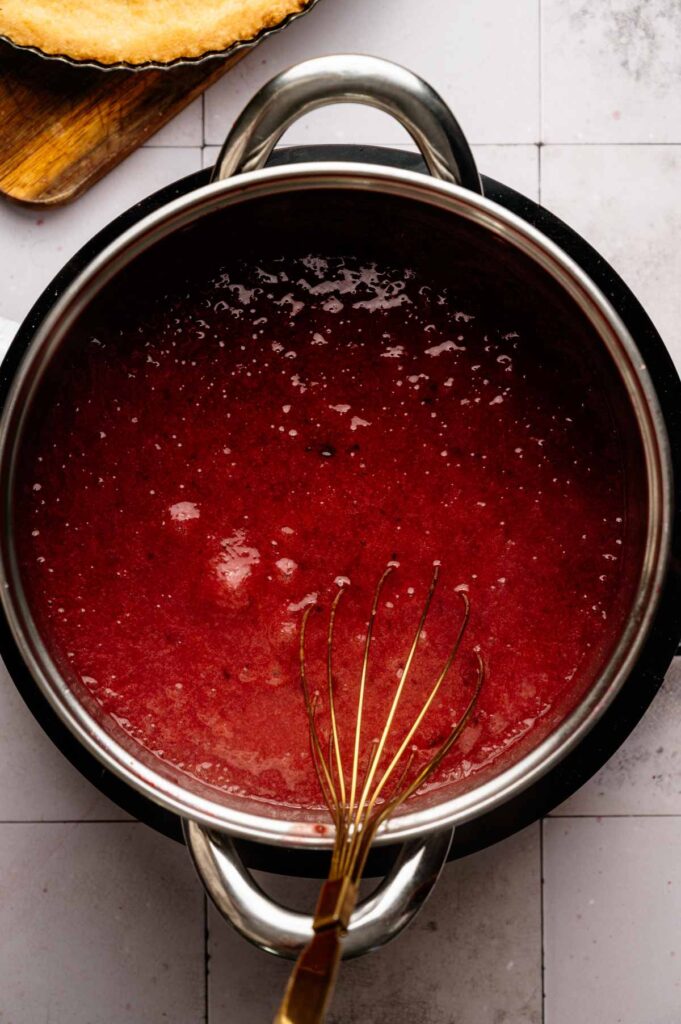 A saucepan filled with a thick red mixture for a Cranberry Curd Tart sits on a stove, with a metal whisk resting inside. A partially visible pie crust is in the upper left corner.