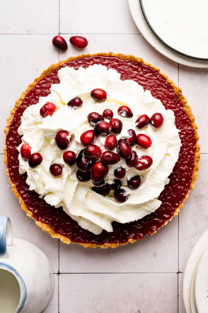 A Cranberry Curd Tart with a layer of whipped cream and fresh cranberries on top, placed on a tiled surface beside stacked plates and a ceramic pitcher.