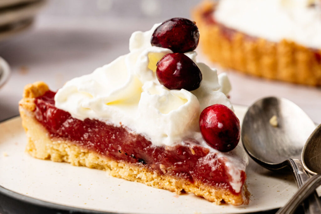 A slice of Cranberry Curd Tart topped with whipped cream and whole cranberries sits on a plate with a spoon beside it.