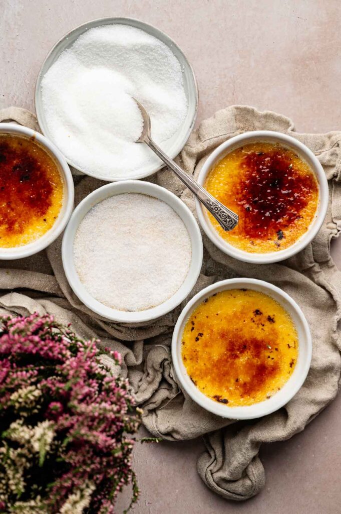 Four ramekins with creamy crème brûlée, one topped with caramelized sugar, and a bowl of granulated sugar with a spoon, arranged on a beige cloth next to a small bouquet of flowers.