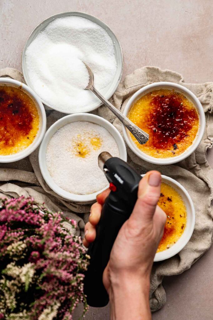 A hand uses a kitchen torch to caramelize sugar on classic Crème Brûlée in white ramekins, with a bowl of granulated sugar and a spoon nearby.