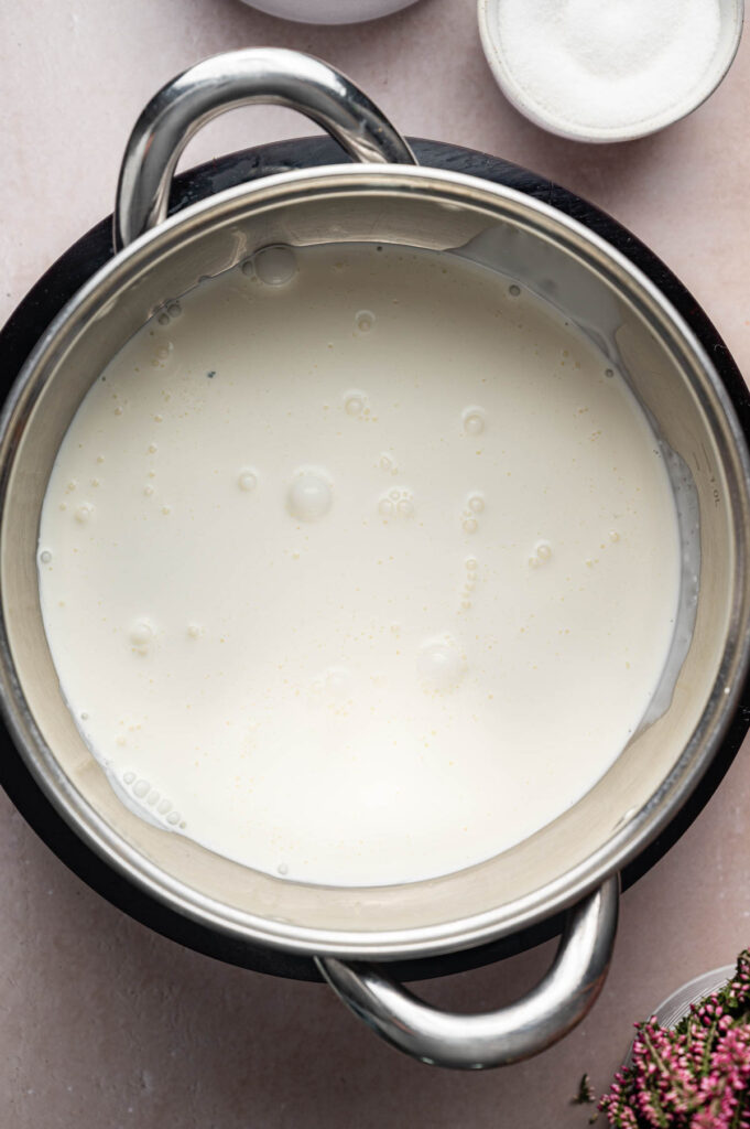 A stainless steel pot filled with milk sits on a light-colored surface, surrounded by small dishes and herbs—perfect ingredients for preparing a creamy Crème Brûlée.