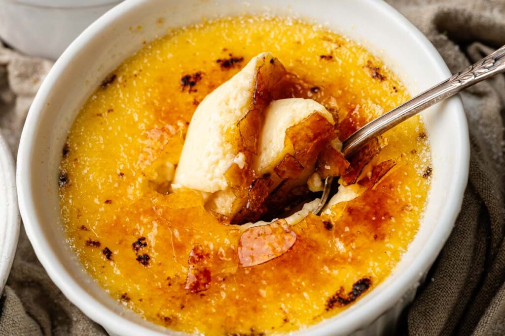 Close-up of classic crème brûlée in a white ramekin, with a spoon breaking through the caramelized sugar crust to reveal the smooth, creamy custard underneath.