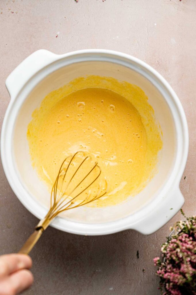 A hand holds a whisk in a white mixing bowl containing a yellow batter, reminiscent of preparing Crème Brûlée. Pink flowers are visible in the bottom right corner on a light-colored surface.