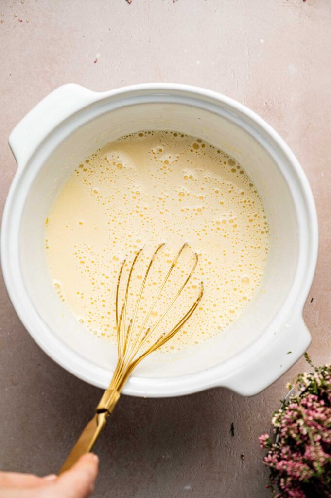 A hand holds a whisk in a white mixing bowl filled with a pale yellow Crème Brûlée mixture, sitting on a light pink surface.