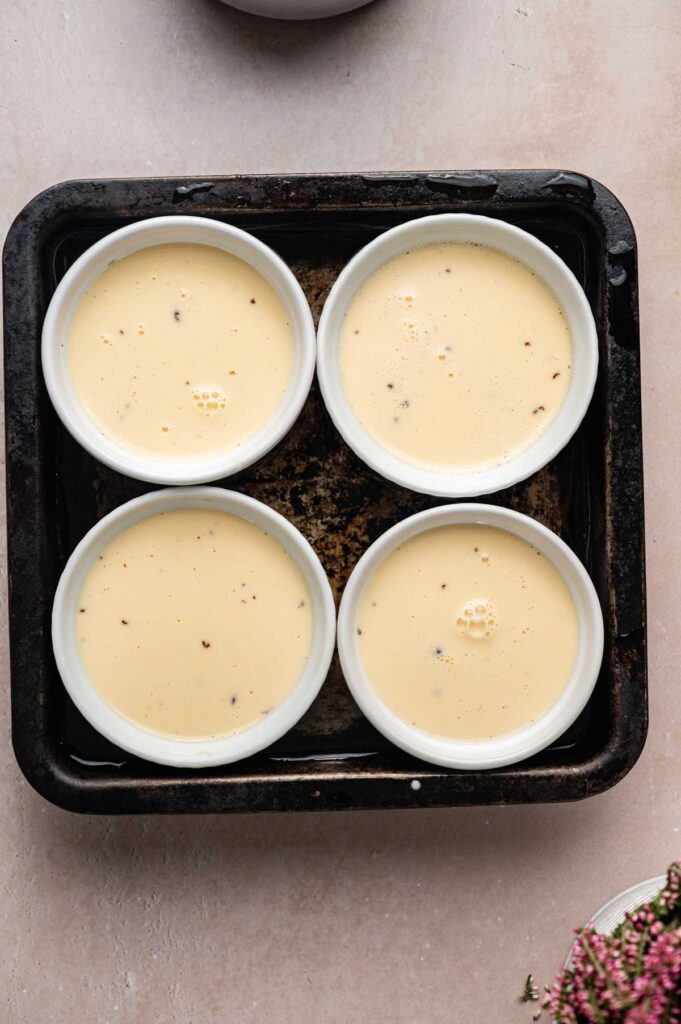 Four white ramekins filled with a creamy Crème Brûlée custard mixture sit in a square metal baking tray, ready for baking.