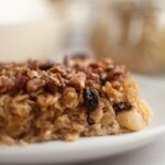 A close-up of a slice of baked oatmeal with nuts and raisins on a white plate, highlighting the hearty texture and wholesome flavors of this classic breakfast treat.