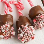Three marshmallows on candy cane sticks are dipped in chocolate and coated with crushed peppermint candies, creating delightful Candy Cane Hot Chocolate Stirrers arranged on a white surface.