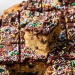 A close-up of a chocolate-frosted Cookie Cake with sprinkles, cut into squares, with one piece lifted above the rest.