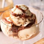 A glass dish with two scoops of creamy No Churn Ice Cream, topped with a drizzle of caramel sauce.